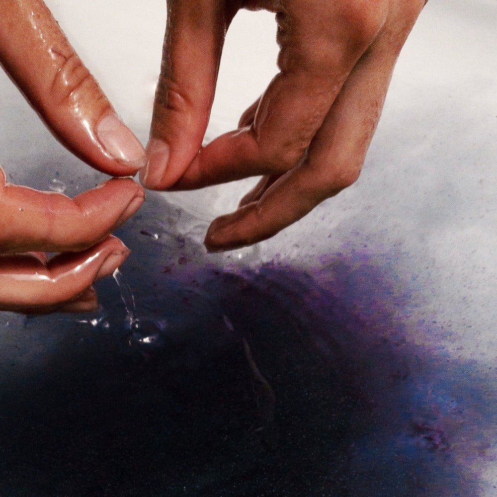 Image of hands dissolving purple herbal bath soak into bath water. 