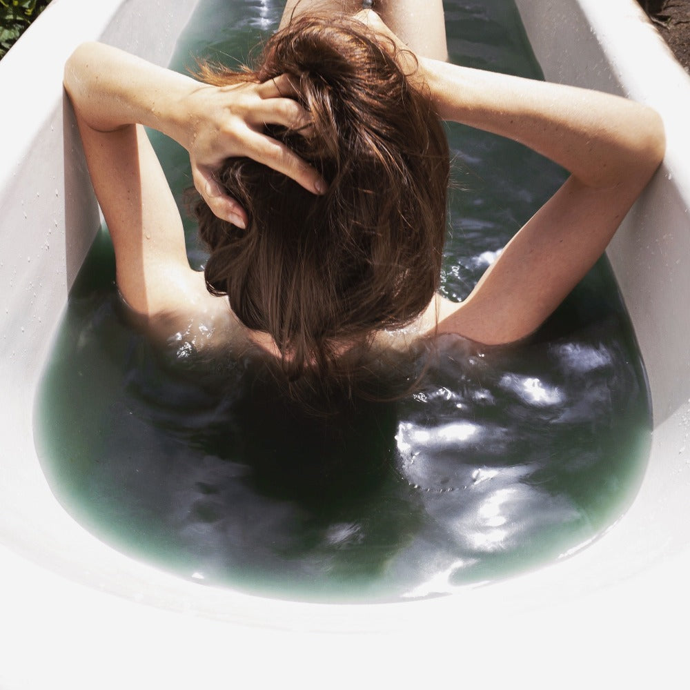 A woman enjoying self-care in a Forgotten Fruit green herbal bath soak.